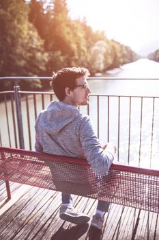 Young man is sitting on bench, enjoying the view over a river