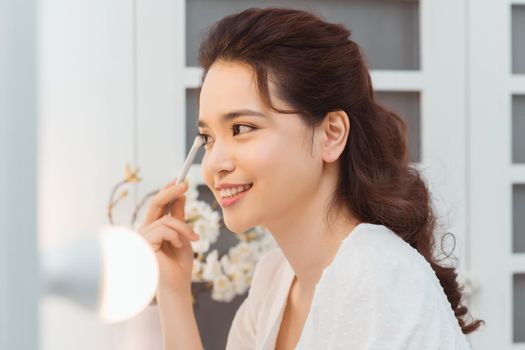 Young stylish woman applying makeup by eye shadow brush in mirror