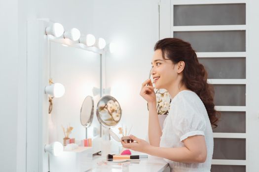 Happy attractive young female applying cosmetics on face near cosmetic table in morning