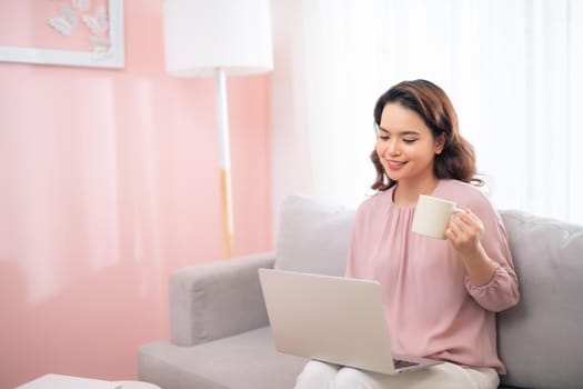 Attractive Asian female lifestyle using the laptop sitting on a couch and drinking coffee