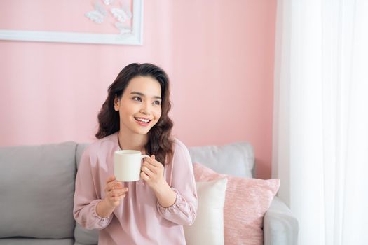 Cheerful young Asian woman holding a mug when sitting on sofa at home.