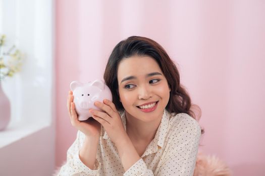 Beautiful young Asian woman holding piggy bank over pink background.