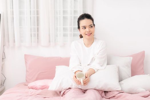 Beautiful happy young woman drinking cup of coffee or tea while lying in bed after waking up in morning. 
