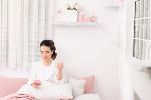 young woman student sitting on bed in her room drinking coffee and using mobile phone