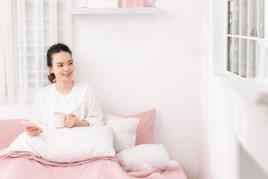 Woman in bed in the bedroom with a mug and a phone in her hands