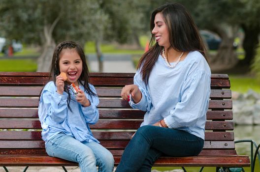 Mother and her little daughter sitting on a bench eating a cookie in the park