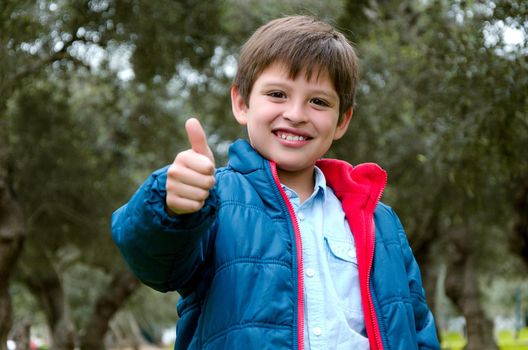 Portrait of a cute blond-haired boy, thumbs up and smile, with green background in the park