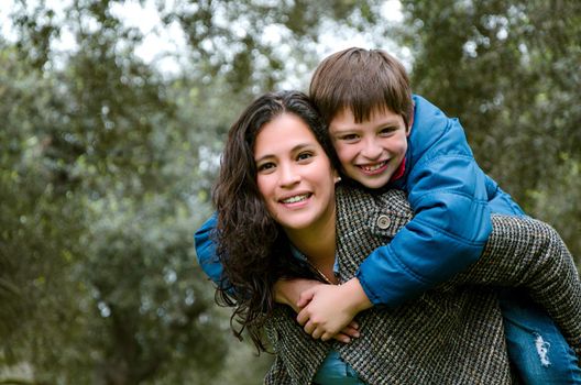 Beautiful mother carrying blond hair son on her back outdoors. Concept of enjoying in family