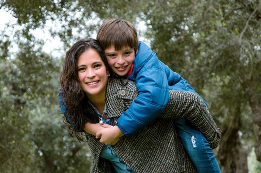 Beautiful mother carrying blond hair son on her back outdoors. Concept of enjoying in family