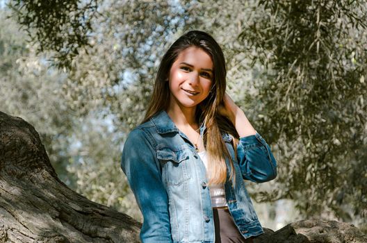 Beautiful long-haired teen woman with brown hair leaning on a tree in the park