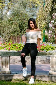 Happy young woman sitting on bench in city park Stylish fashion model wearing ripped jeans and pullover