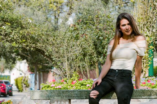 Happy young woman sitting on bench in city park Stylish fashion model wearing ripped jeans and pullover