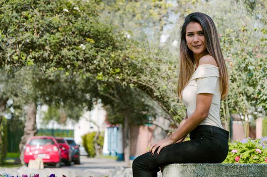 Happy young woman sitting on bench in city park Stylish fashion model wearing ripped jeans and pullover