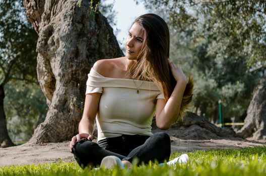 Beautiful brunette woman touching her long hair. She sits in a park on green grass. The image is toned.