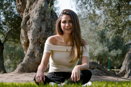 Stylish cheerful brunette sitting under a tree in a park on a sunny day