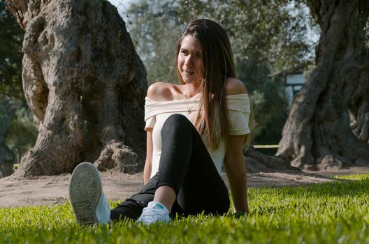 Stylish cheerful brunette sitting under a tree in a park on a sunny day