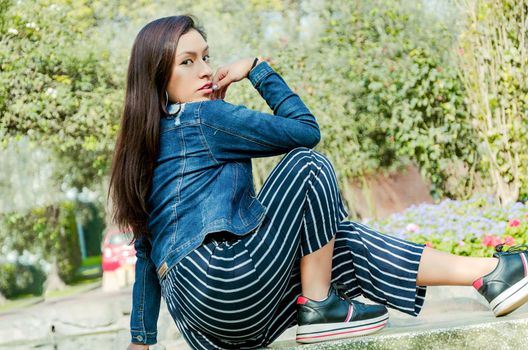 Outdoors lifestyle fashion portrait of pretty fashionable woman sitting on the marble bench. With an elegant wide jacket of jeans and black pants