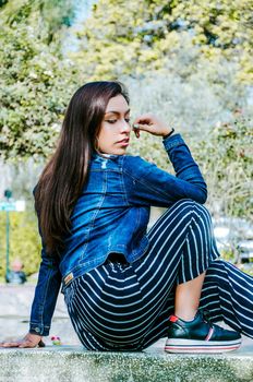 Outdoors lifestyle fashion portrait of pretty fashionable woman sitting on the marble bench. With an elegant wide jacket of jeans and black pants