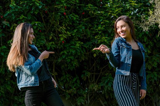 Cheerful friends are gossiping in the park. The girls are pointing with their finger and laughing. They wear fashionable clothes