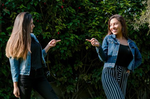 Cheerful friends are gossiping in the park. The girls are pointing with their finger and laughing. They wear fashionable clothes