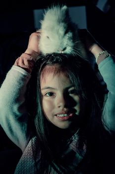 Happy little girl with her pet bunny on her head on dark background