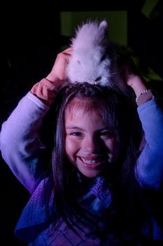 Happy little girl with her pet bunny on her head on dark background