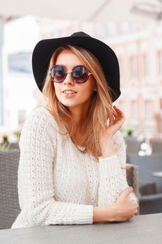 Portrait of a young pretty woman in a black hat and round sunglasses. Beautiful girl sits in summer cafe.