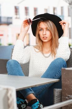 Portrait of a young pretty woman in a black hat and round sunglasses. Beautiful girl sits in summer cafe.