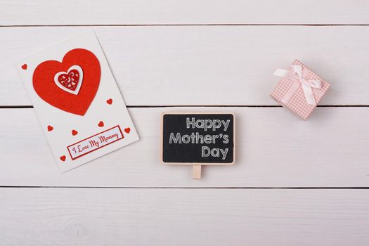 Blackboard with happy mathers day tag, a gift and a card with red hearts on a white table.