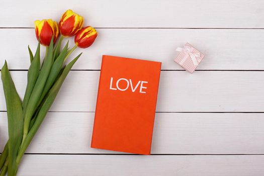 The red book Love lies on a white table. Flowers tulips and gift with a bow on the table.
