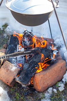 Over the fire hangs a pot in which to cook food. On a hook on a tripod, steam comes out of the pan. Winter Camping outdoor cooking