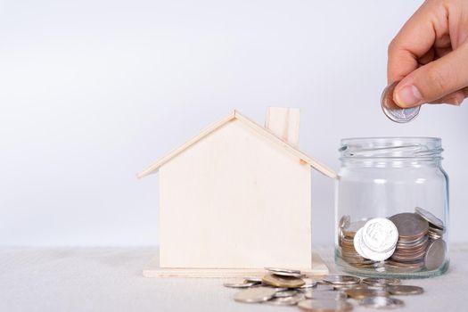 Hand put money coin into jar with wooden house isolated grey background. Saving money and investment concept.