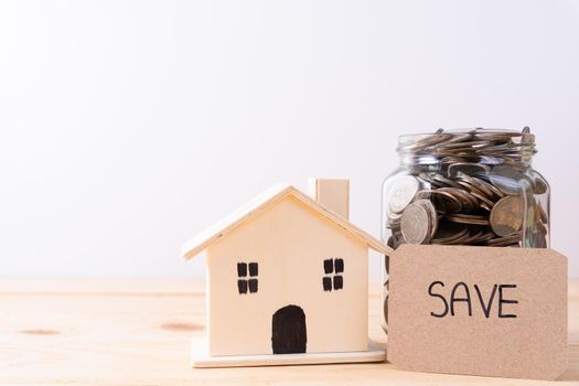 Jar full of coins with save word paper and wooden house on wooden table isolated grey background. Saving money and investment concept.