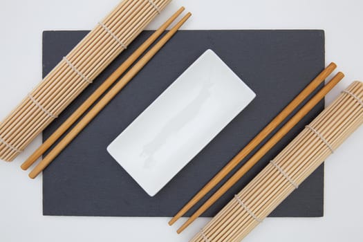Rectangular slate plate with chopsticks, ceramic plate, bamboo mat for sushi on the white table.