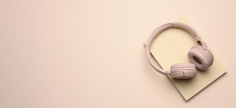 beige wireless headphones and a closed notepad on a beige background, banner. Top view and place for an inscription