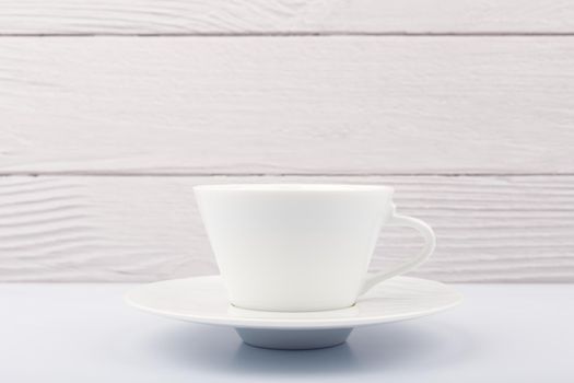 Close up of white ceramic coffee or tea cup with saucer on white table against white blurred wooden background