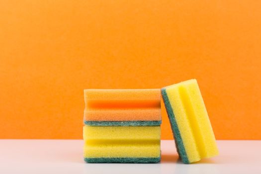 Minimalistic still life with yellow and orange sponges for house cleaning and dish washing on white table against orange background. Concept of cleaning tools for home