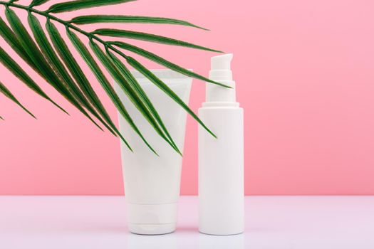 Two white unbranded cream tubes on white table against pink background with palm leaf. Concept of skin care and beauty treatment for smooth, young looking skin