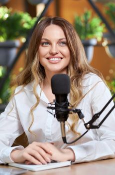 Close up portrait of smiling cute woman with long hair in white shirt communicating with followers online with modern black microphone. Concept of process talking blogger and subscribers online. 