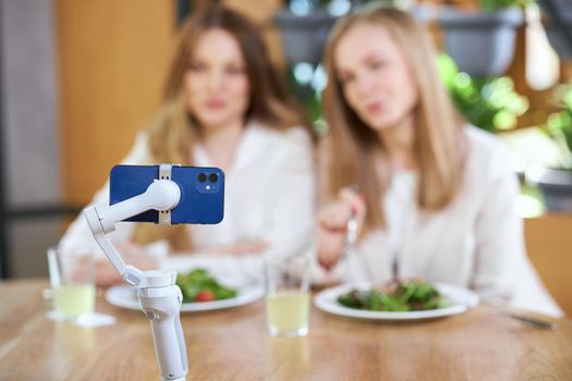 Front view of two beautiful women eating on camera and communicating with followers in live broadcast. Concept of talking with subscribers online in cafe with good mood. 