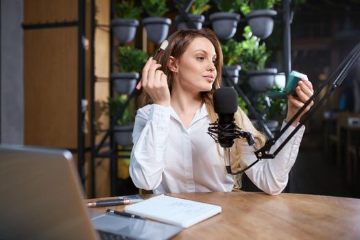 Front view portrait of attractive young woman preparing for interview and doing makeup with cosmetic. Concept of sitting at table near modern black microphone and laptop.