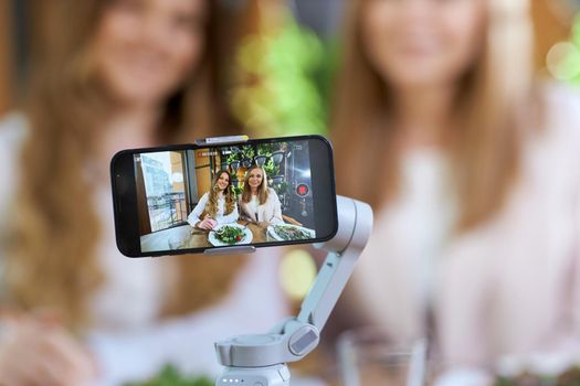 Close up of two beautiful young blogger women posing on phone camera. Concept of process photo shoot on modern phone in cafe with delicious food and with good mood.