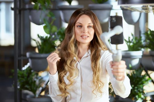 Front view of attractive woman with long hair in white shirt holding selfie stick and communicating online with followers or friends on phone. Concept of process doing selfie. 