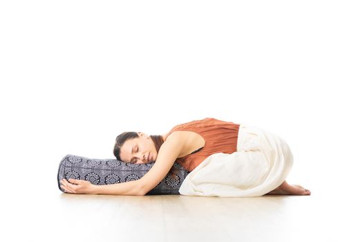 Restorative yoga with a bolster. Young sporty attractive woman in bright white yoga studio, lying on bolster cushion, stretching and relaxing during restorative yoga. Healthy active lifestyle.