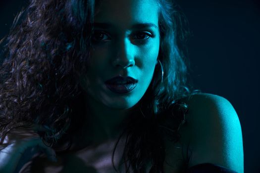 Portrait of glamour brunette model with beautiful curly hair and trendy accessories in dark blue light on photo studio. Concept of process shooting with special atmosphere.