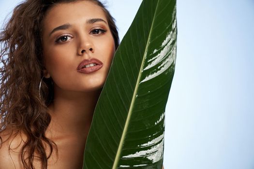 Portrait of beautiful model with curly hair with big green leaf on light blue background. Concept of shooting with green plants for fashion magazine on professional studio.