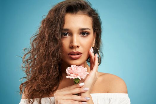 Close up portrait of young beautiful model with curly hair and holding pretty pink flower on blue background. Concept of modern professional shooting in photo studio. 