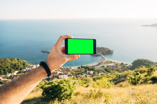 Man holds horizontally in his hand a smartphone with a blank screen against the background of Sveti Stefan island. High quality photo
