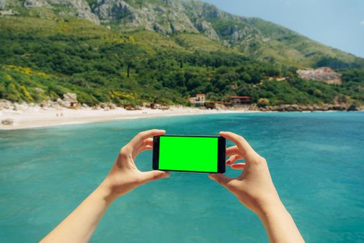Man's hands hold a smartphone with a blank screen horizontally against a background of greenery and blue sea. High quality photo