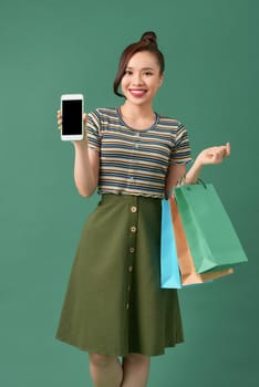 smiling young woman showing blank screen smrtphone and holding bags